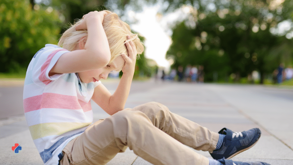 image of young boy sitting on ground outside,, holding is head demonstrating one of the key signs of a concussion